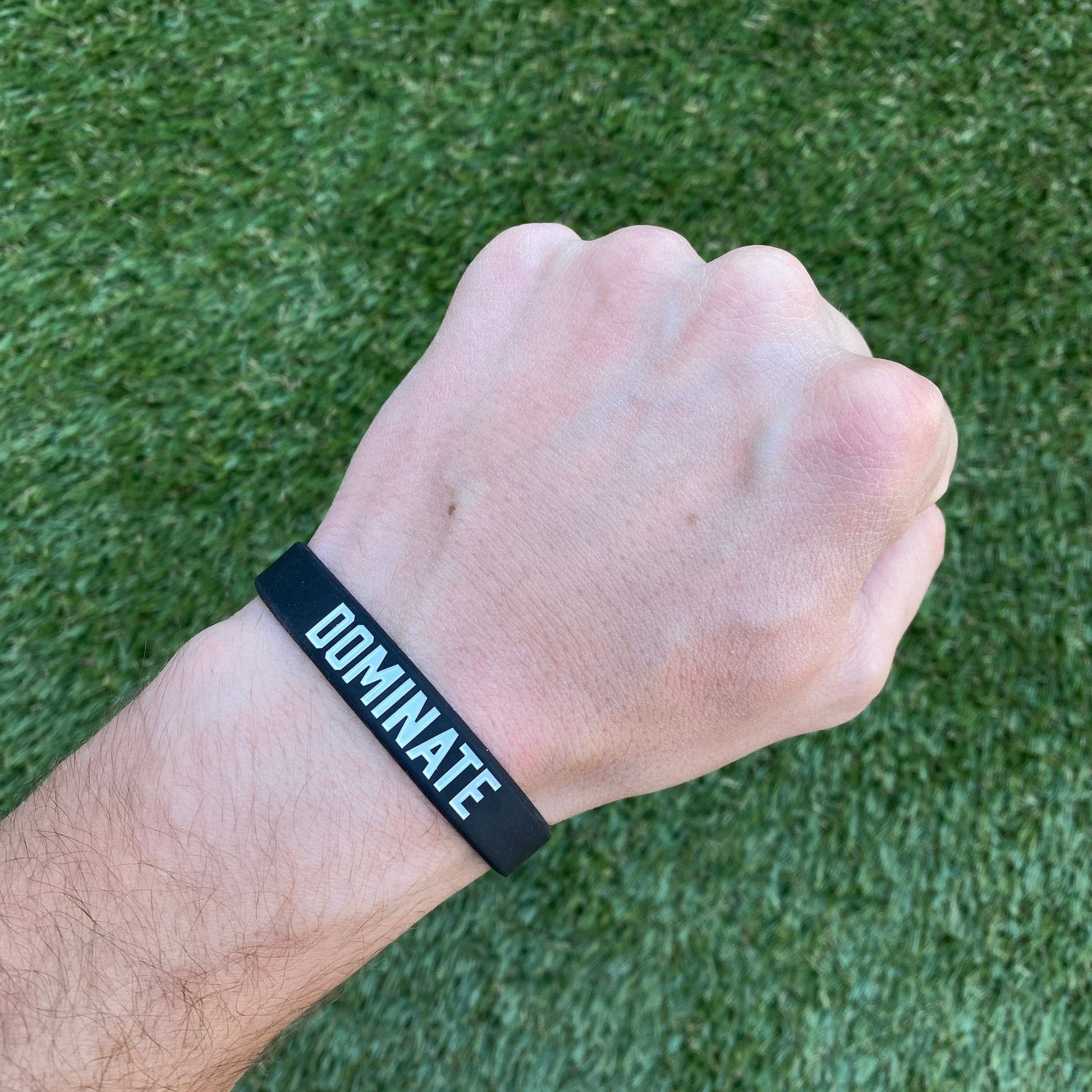 Hand wearing a black wristband with 'DOMINATE' text on a grass background