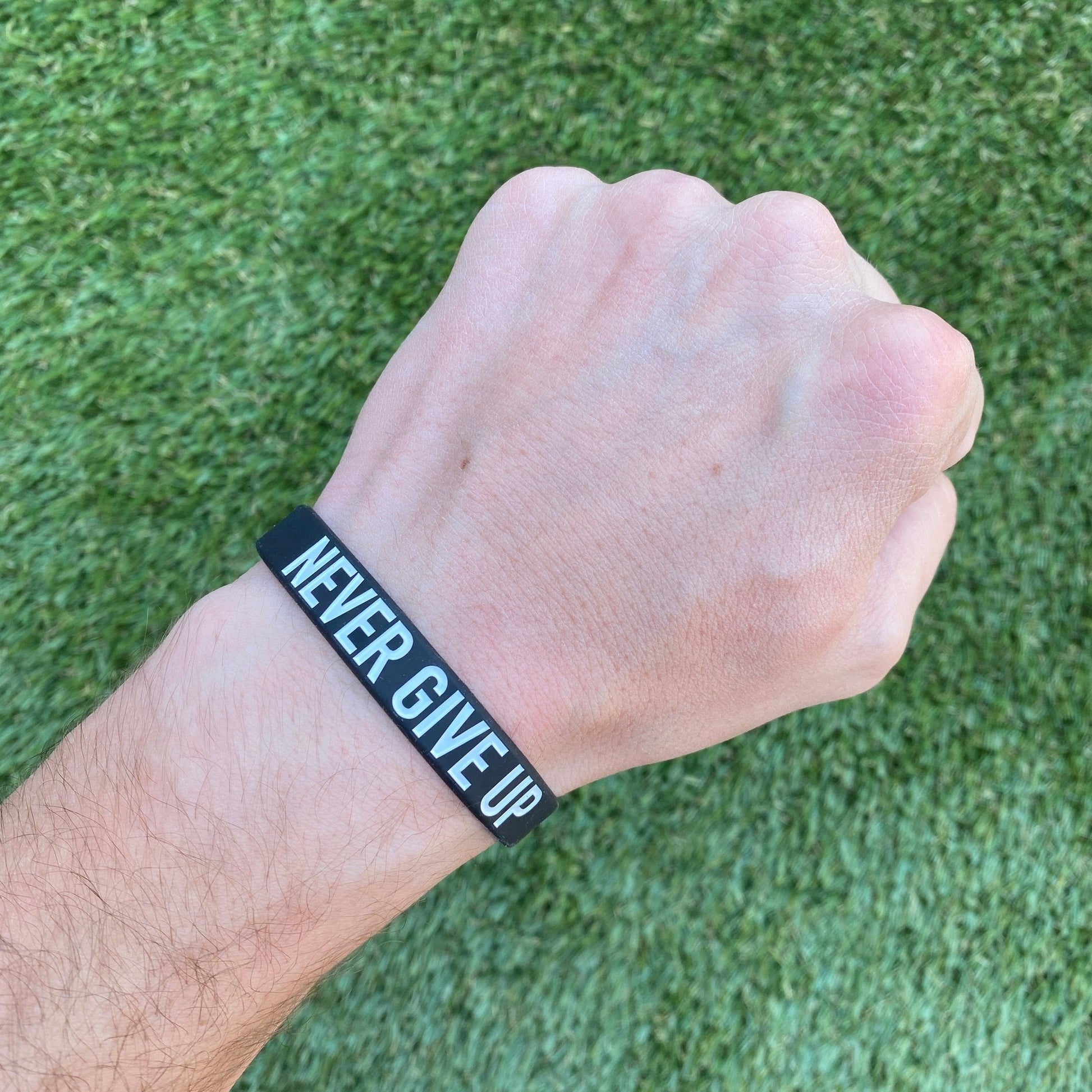 Hand wearing a black wristband with 'NEVER GIVE UP' text on a grass background