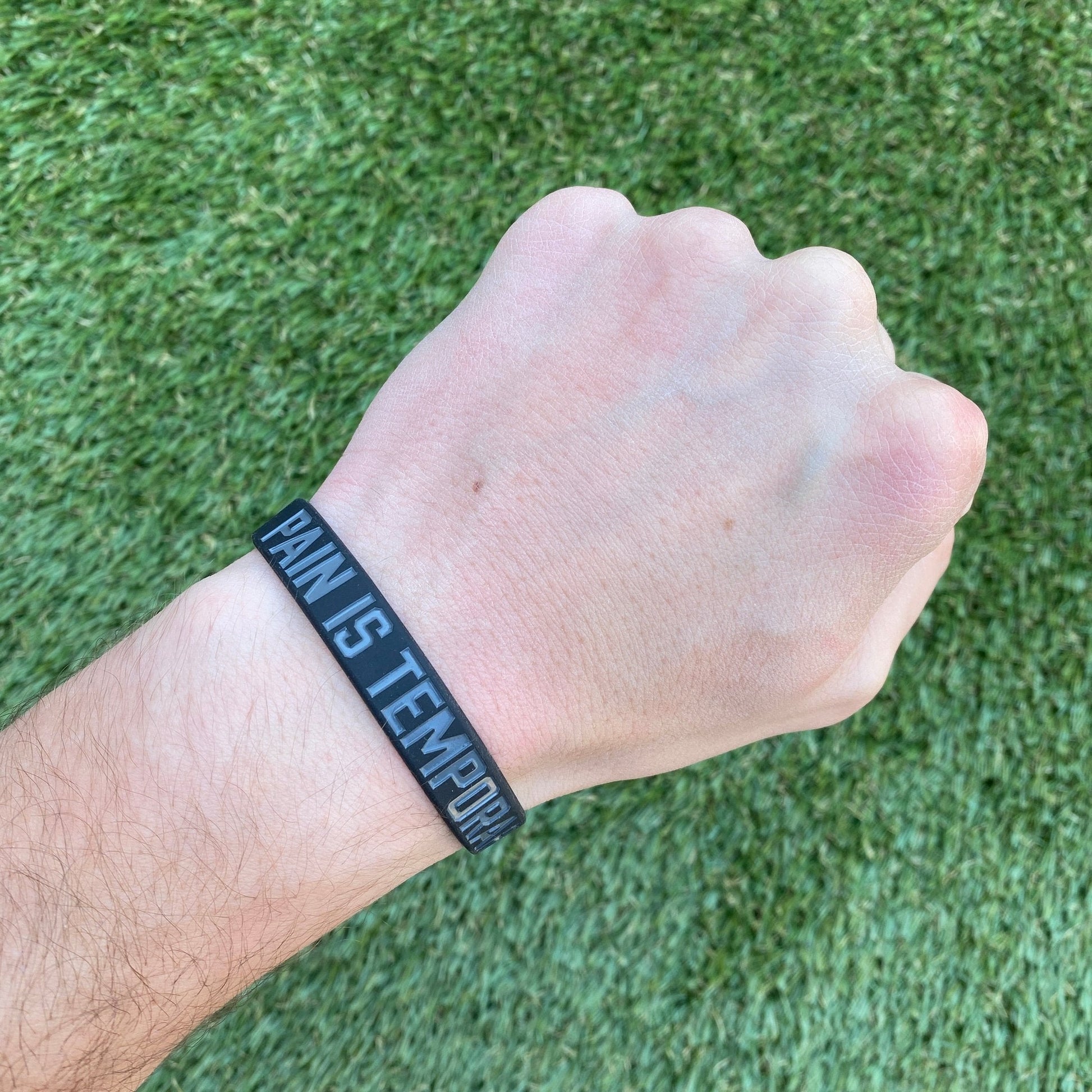 Hand wearing a black wristband with 'PAIN IS TEMPORARY' text on a grass background