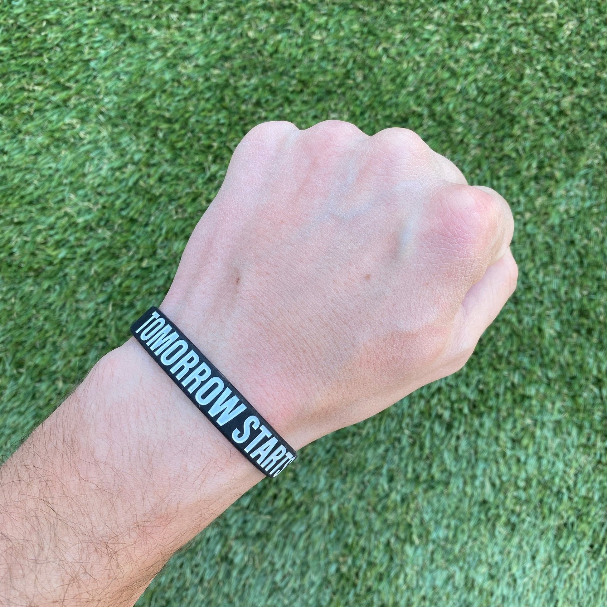 Hand wearing a black wristband with 'TOMORROW STARTS HERE' text on a grass background