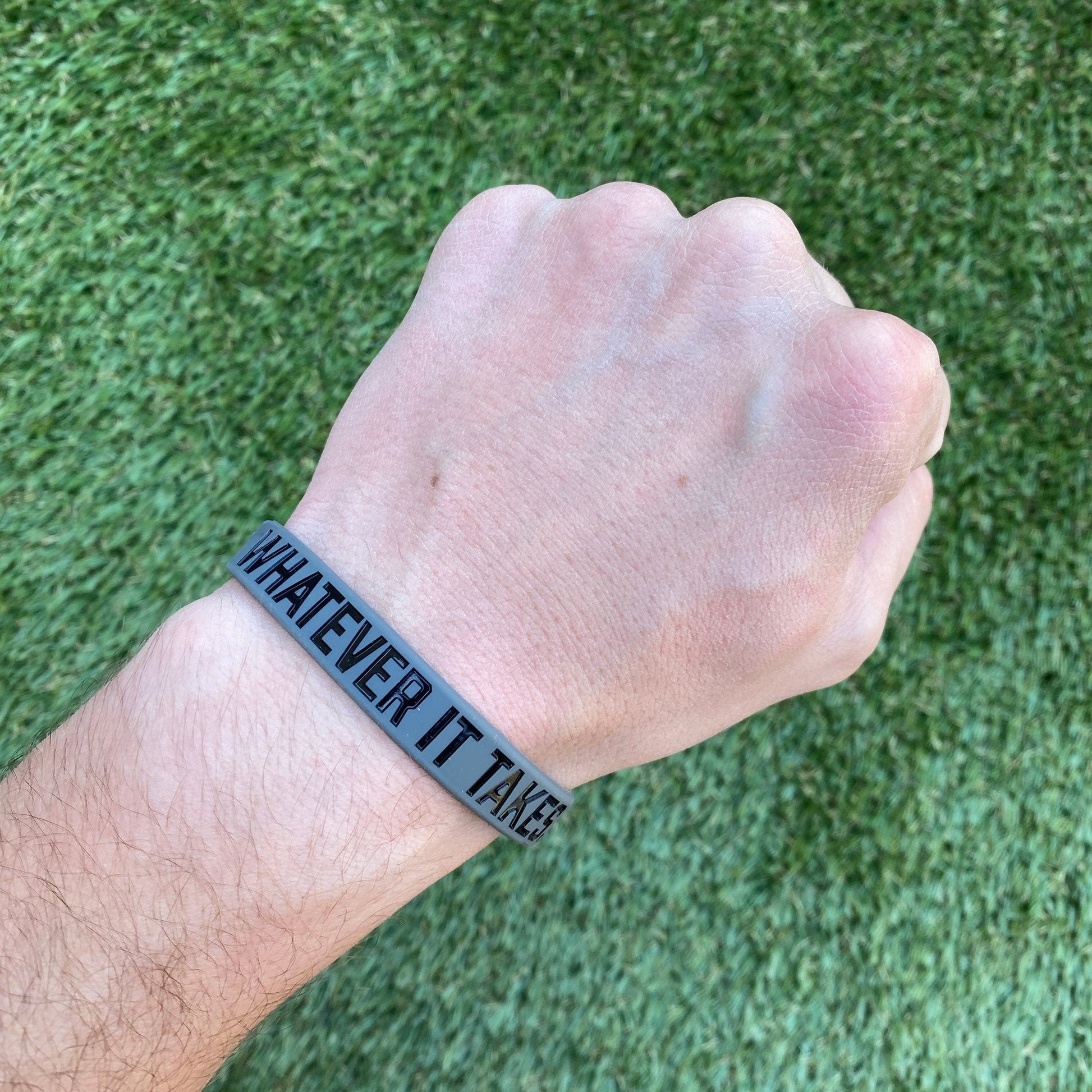 Hand wearing a wristband with 'Whatever It Takes' text on a grass background