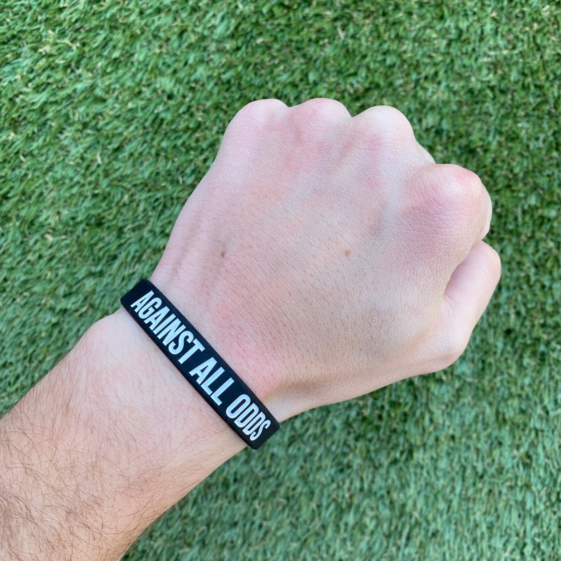 Hand wearing a black wristband with 'Against All Odds' text on a grass background