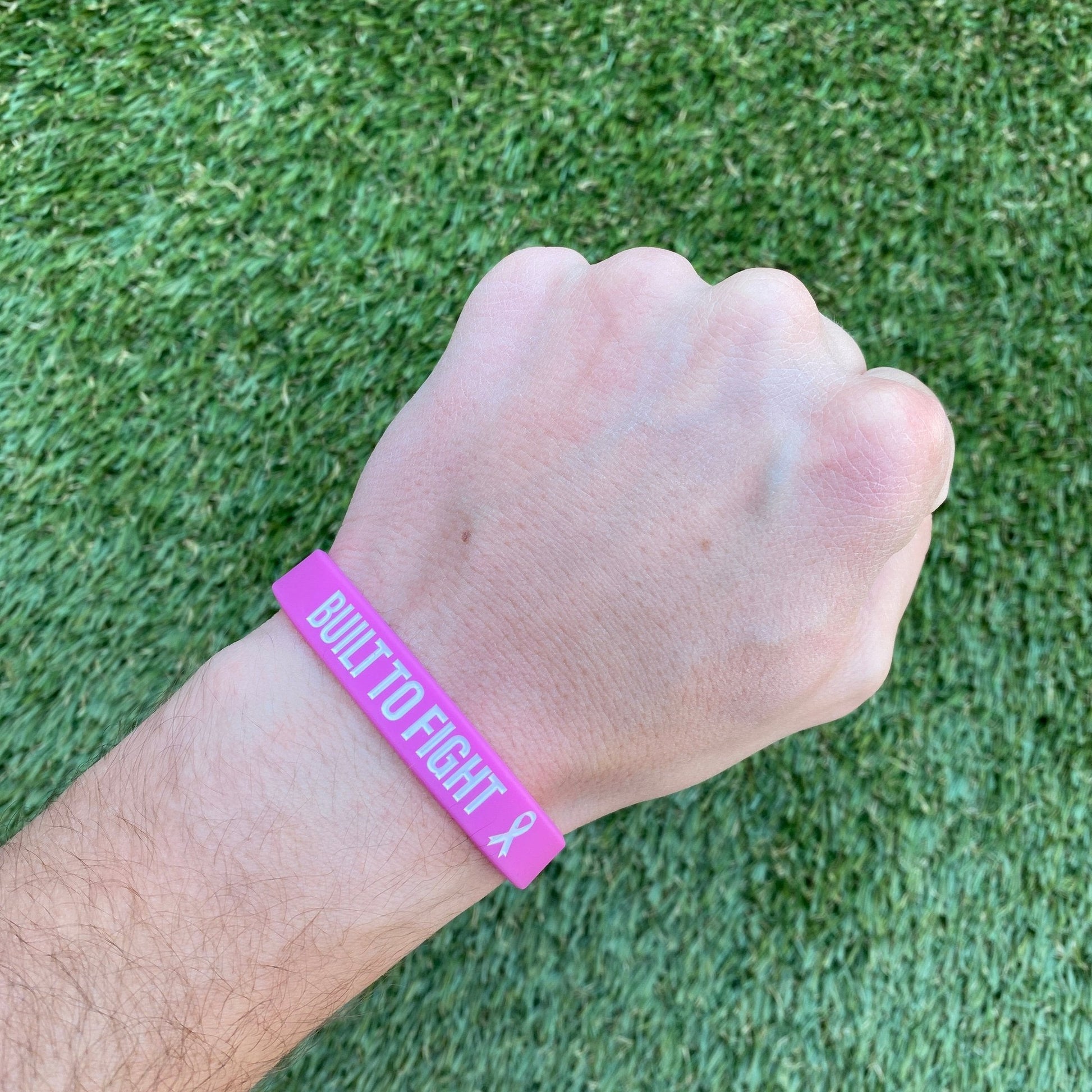 Hand wearing a pink wristband with 'BUILT TO FIGHT' text on grass background