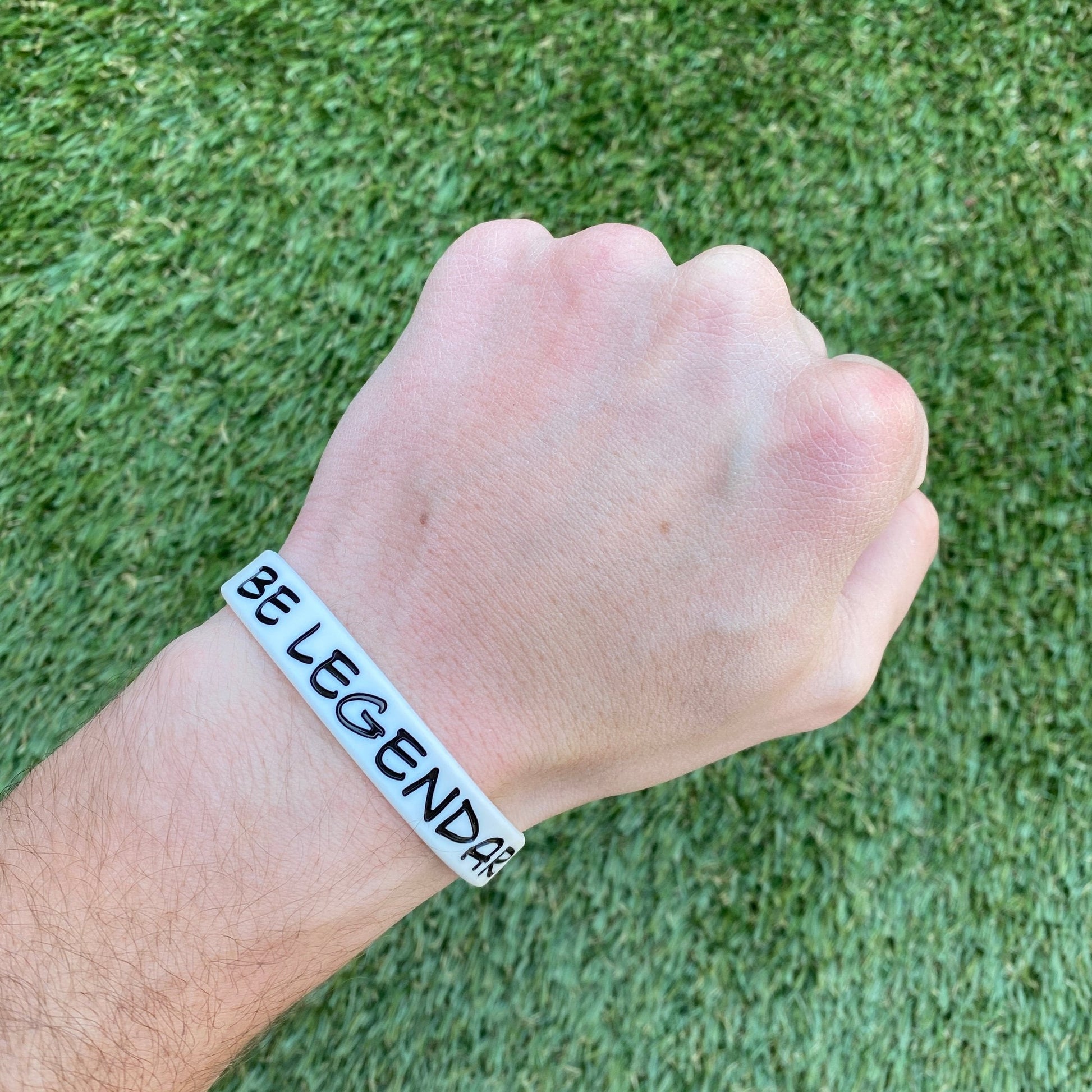 Hand wearing a bracelet with 'BE LEGENDARY' text on a grass background
