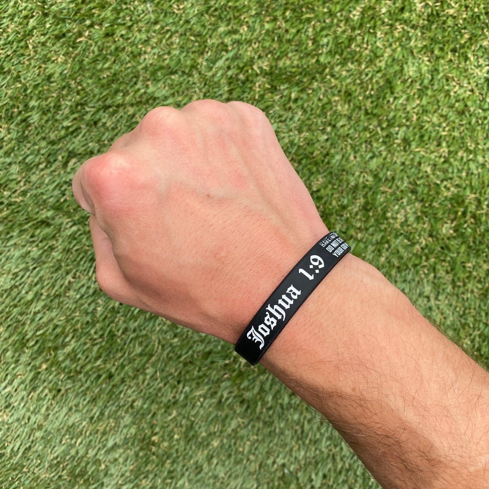 Black wristband with 'Joshua 1:9' text on a wrist against a grass background