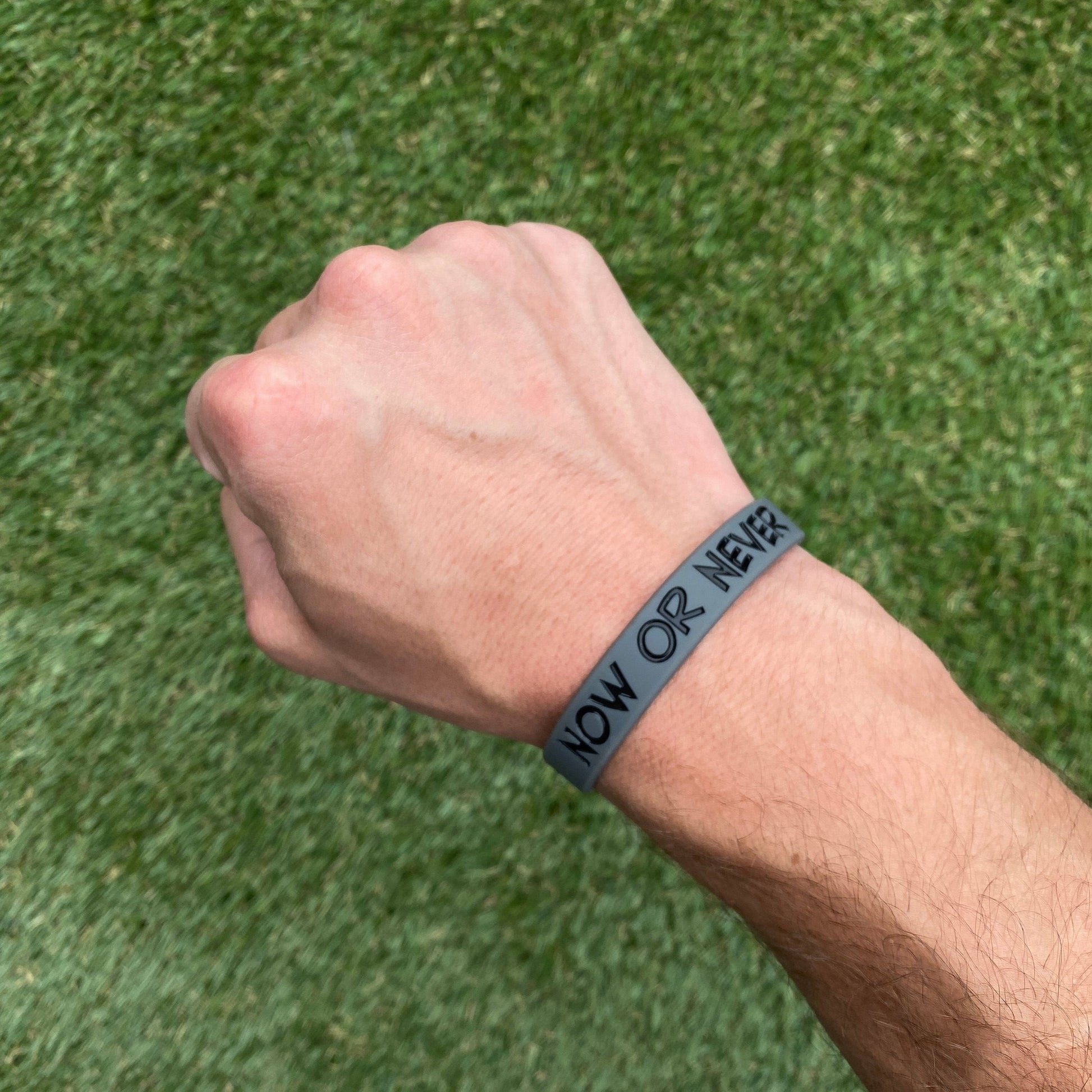Wrist wearing a 'NOW OR NEVER' bracelet against a grass background