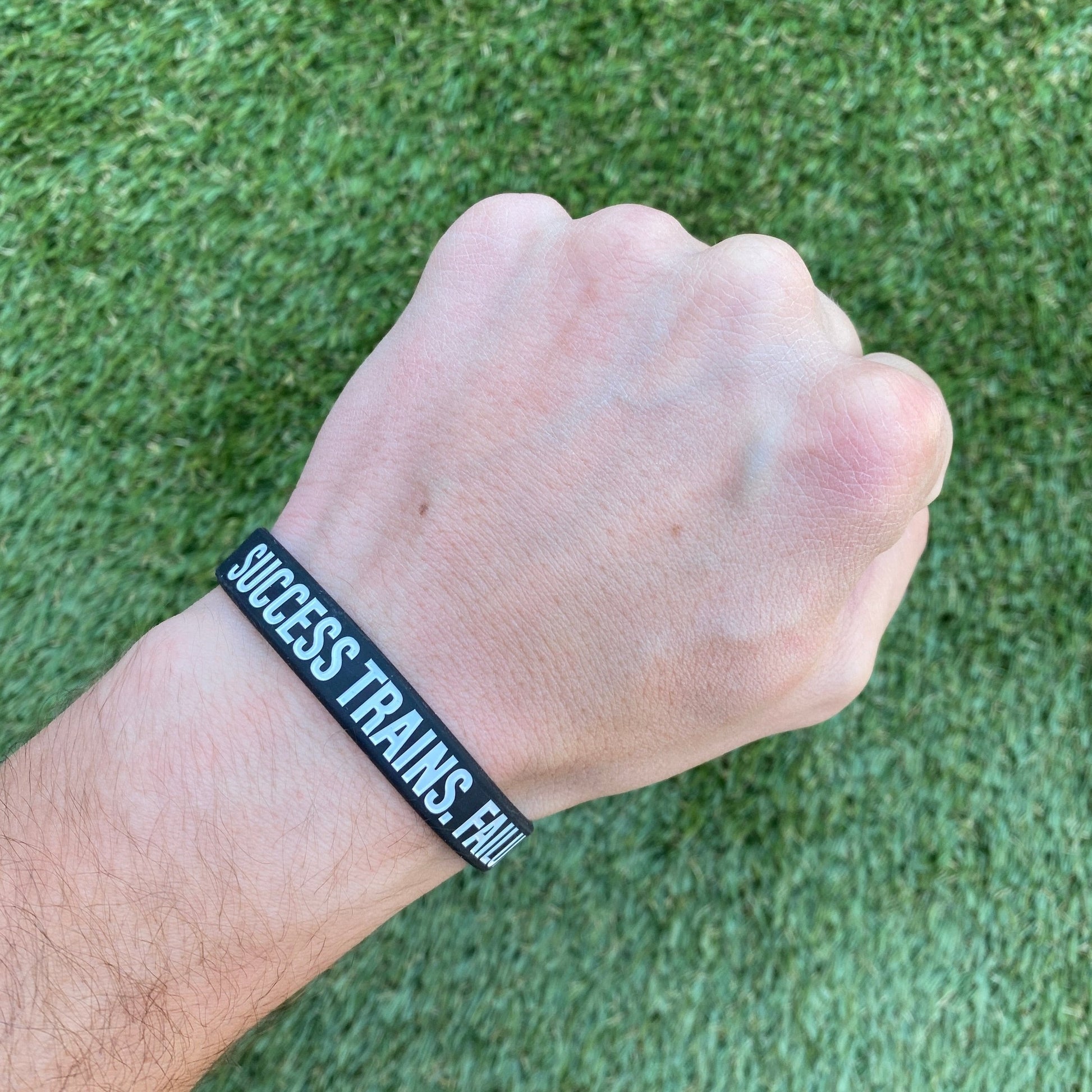 Hand wearing a wristband with 'SUCCESS TRAINS, FAIL' on a grass background