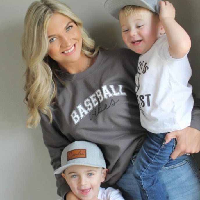 Woman wearing a 'Baseball Vibes' sweatshirt with two children, one wearing a cap, on a plain background.