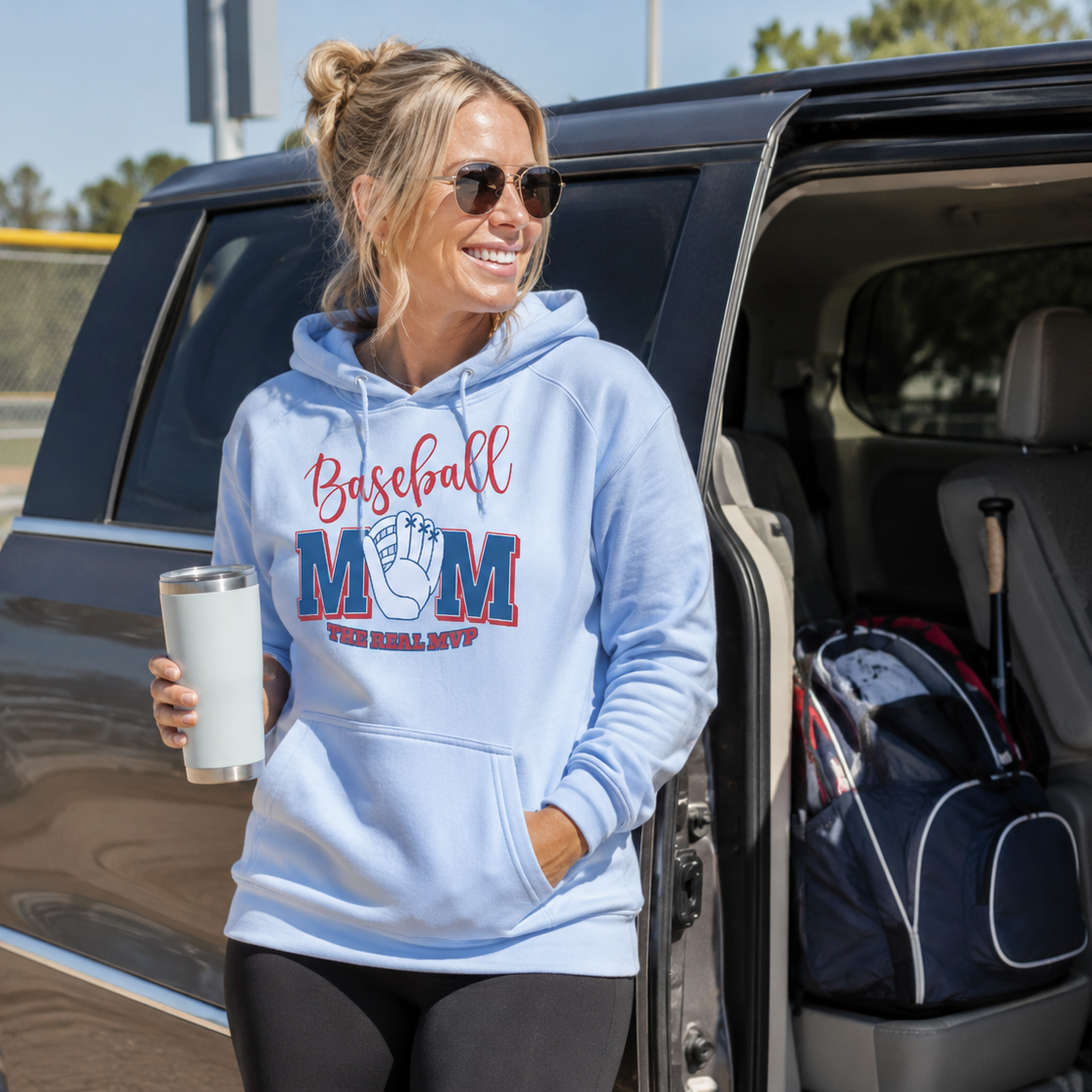 Woman in a 'Baseball Mom' hoodie standing next to an open car door, holding a tumbler.
