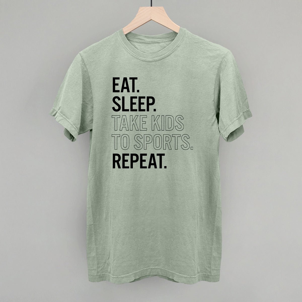 Green comfort colors t-shirt with text 'Eat. Sleep. Take kids to sports. Repeat.' on a gray background