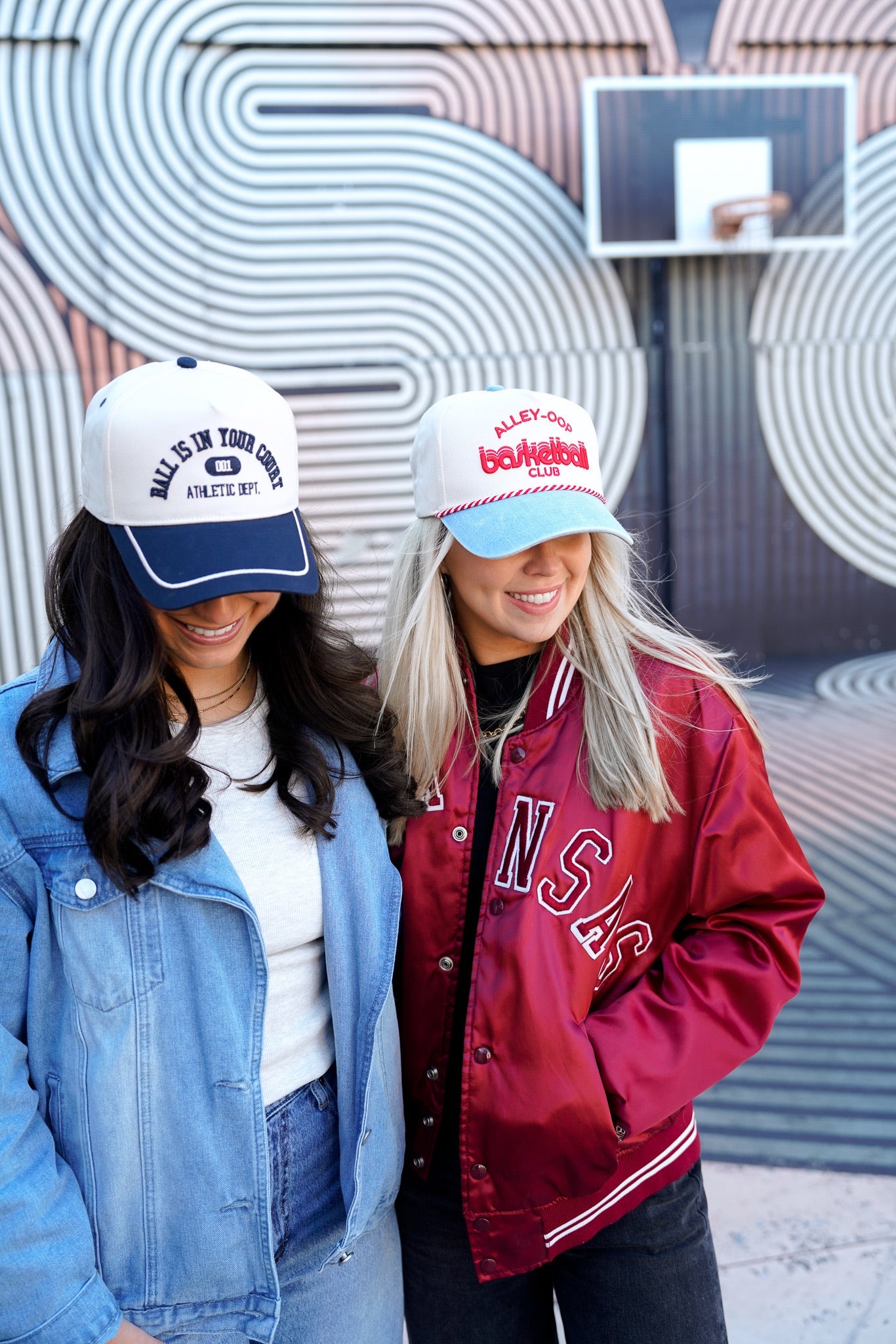 Collection of Two women wearing baseball caps and jackets standing in front of a wall with circular patterns. in a gallery layout