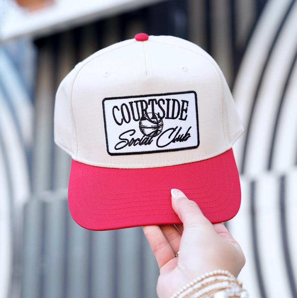 Hand holding a red and white cap with 'Courtside Social Club' logo against a blurred outdoor background.