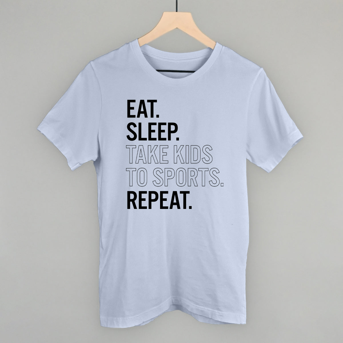 Light blue comfort color t-shirt with text 'EAT. SLEEP. TAKE KIDS TO SPORTS. REPEAT.' on a hanger against a gray background