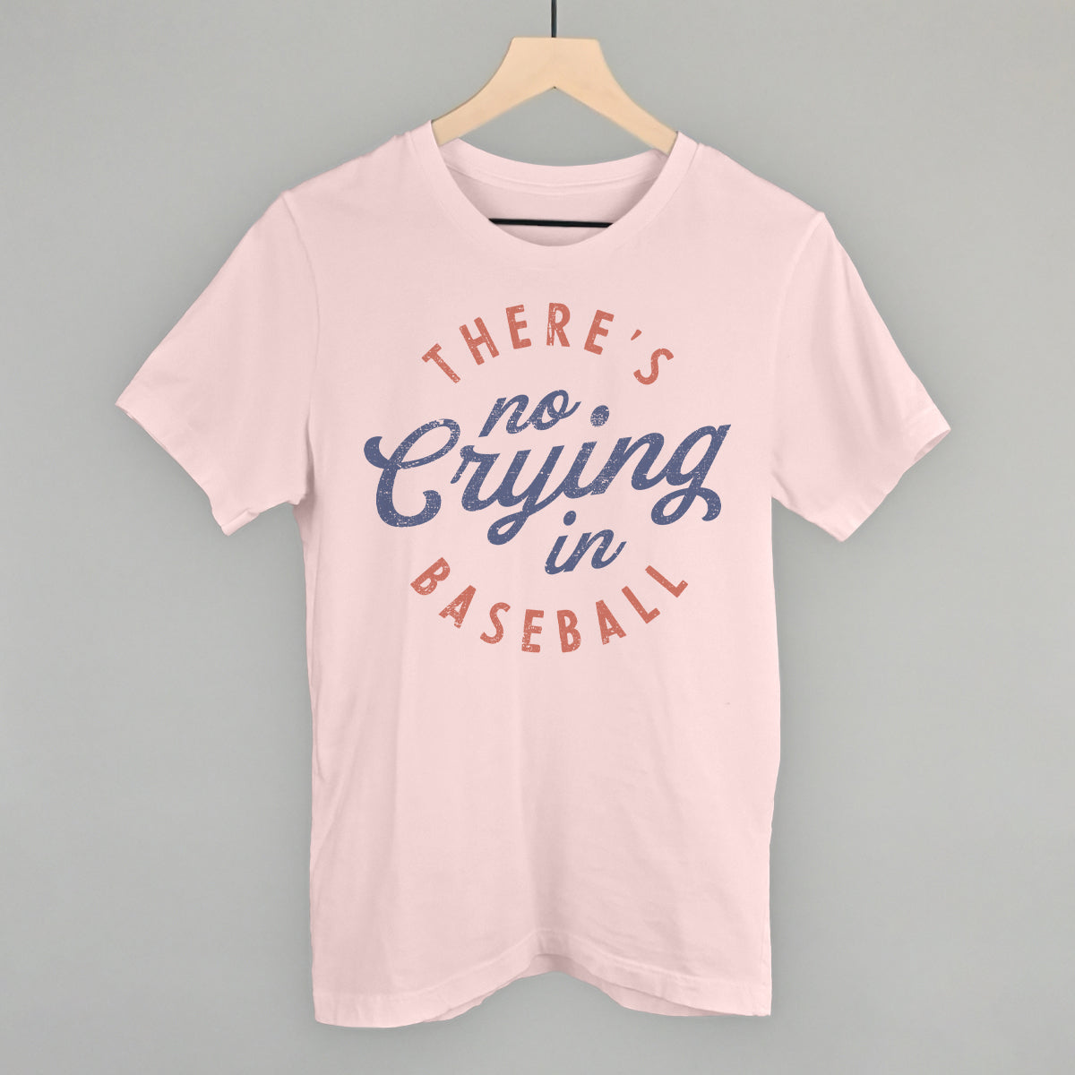 Pink t-shirt with 'There's no crying in baseball' text on a hanger against a gray background