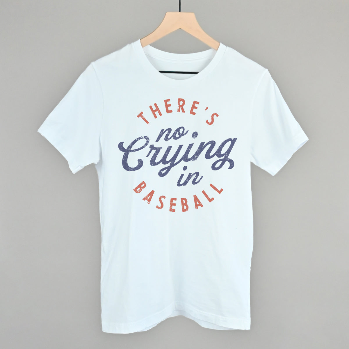 White t-shirt with 'There's no crying in baseball' text on a hanger against a gray background
