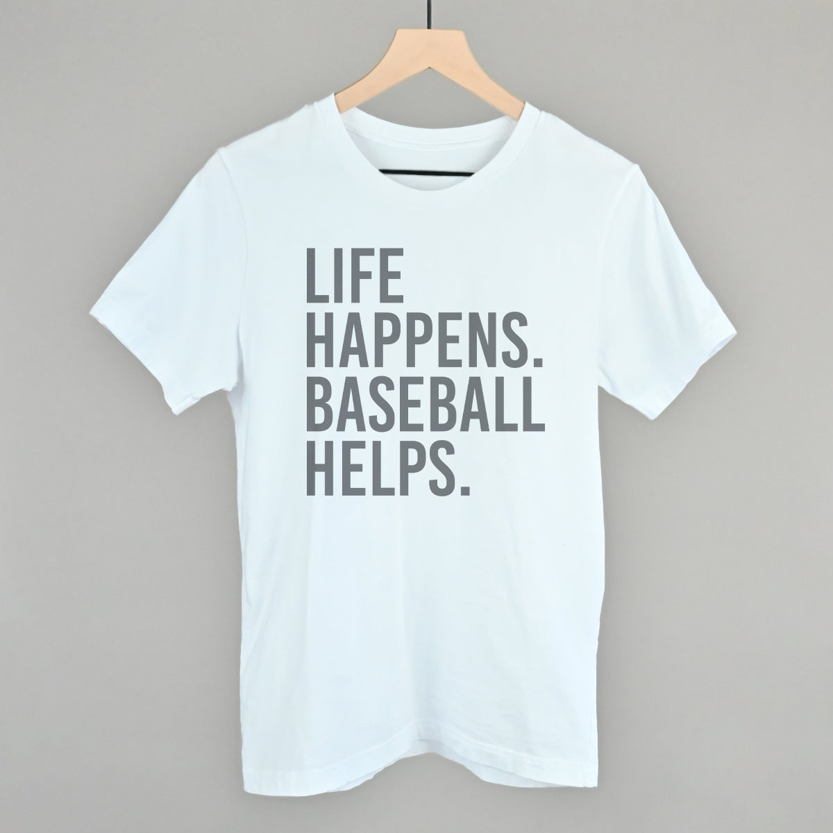 White t-shirt with 'Life happens. Baseball helps.' text on a hanger against a gray background
