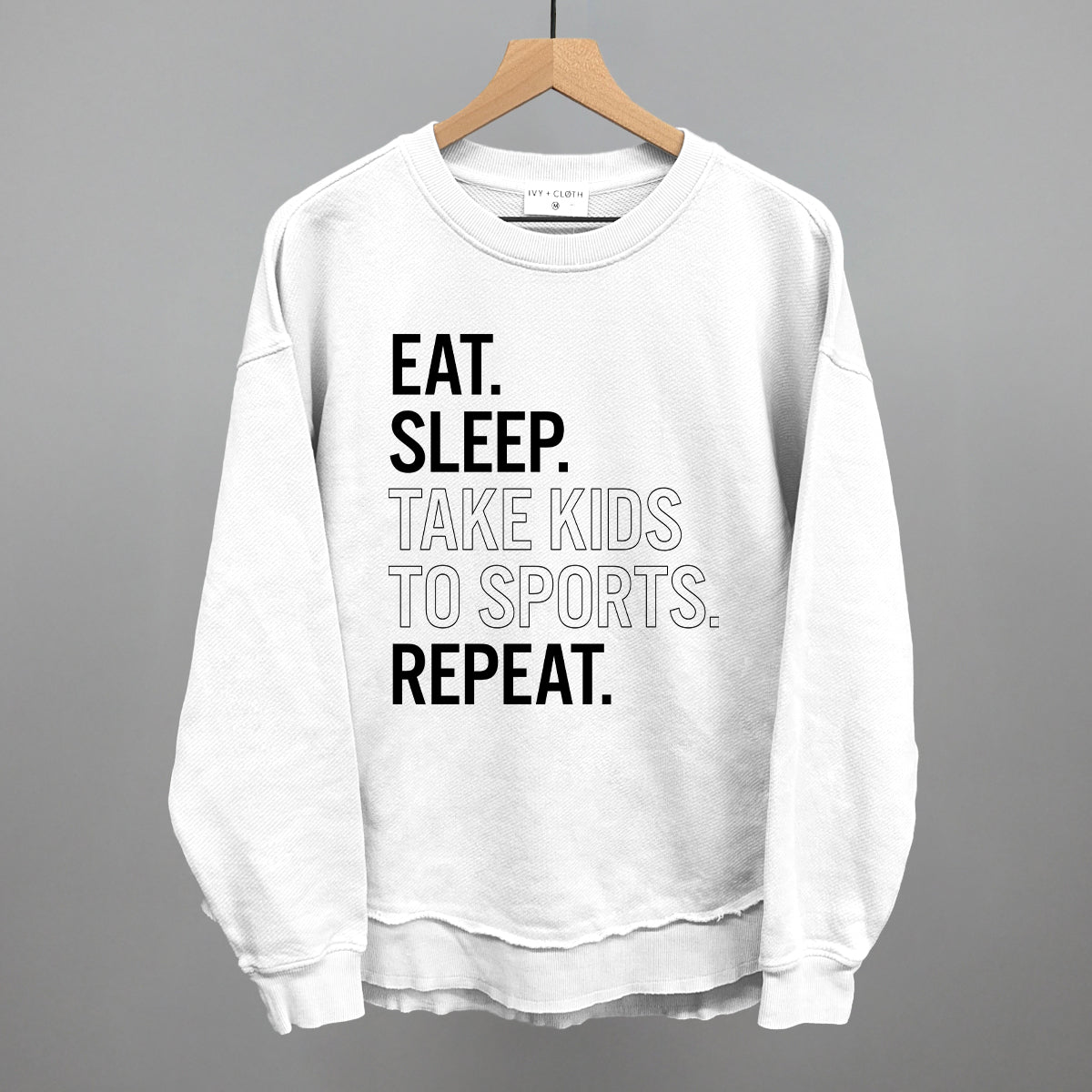 White sweatshirt with text 'EAT. SLEEP. TAKE KIDS TO SPORTS. REPEAT.' on a hanger against a gray background