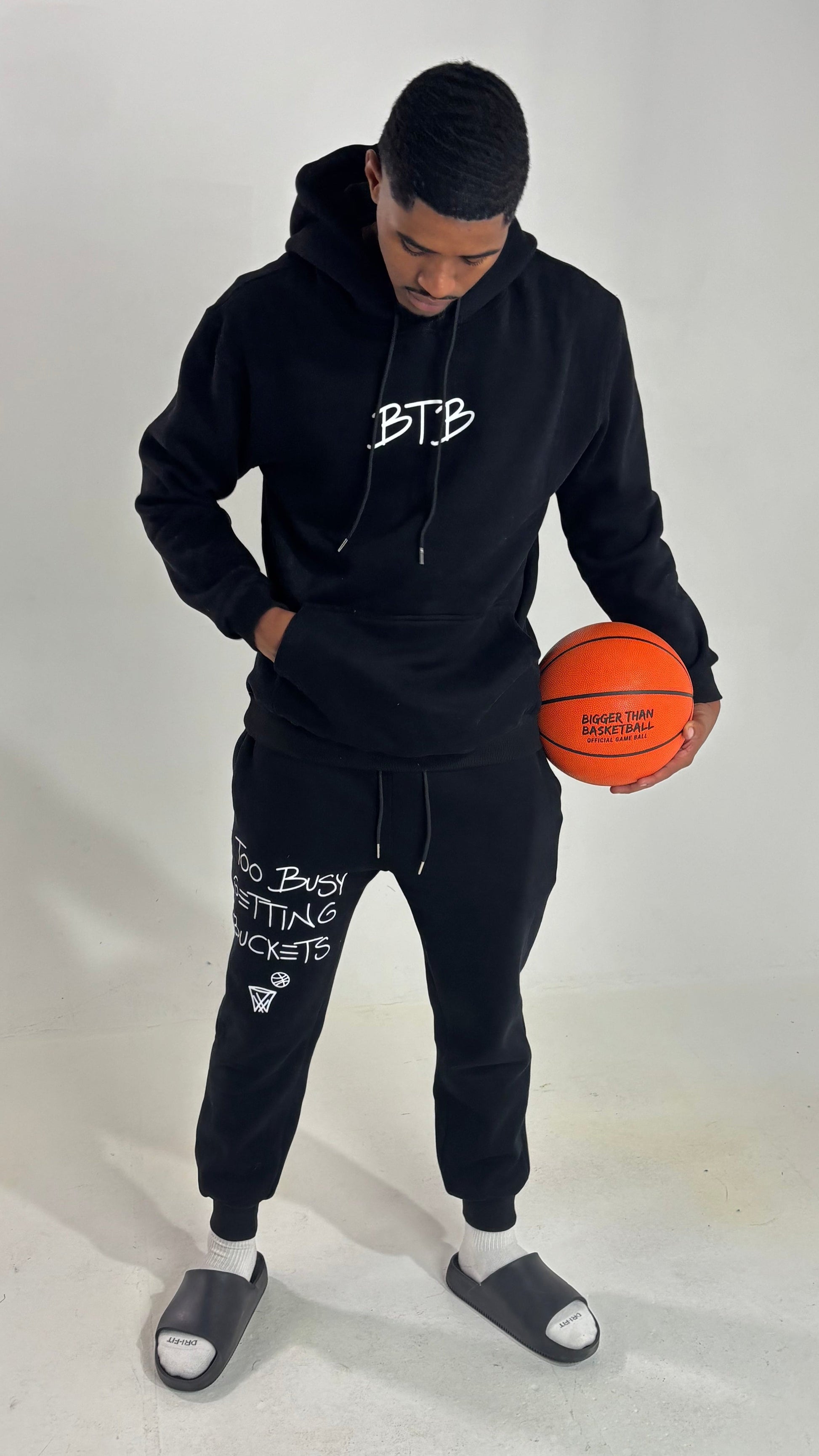 Person wearing a black hoodie and jogger set holding an orange basketball on a white background