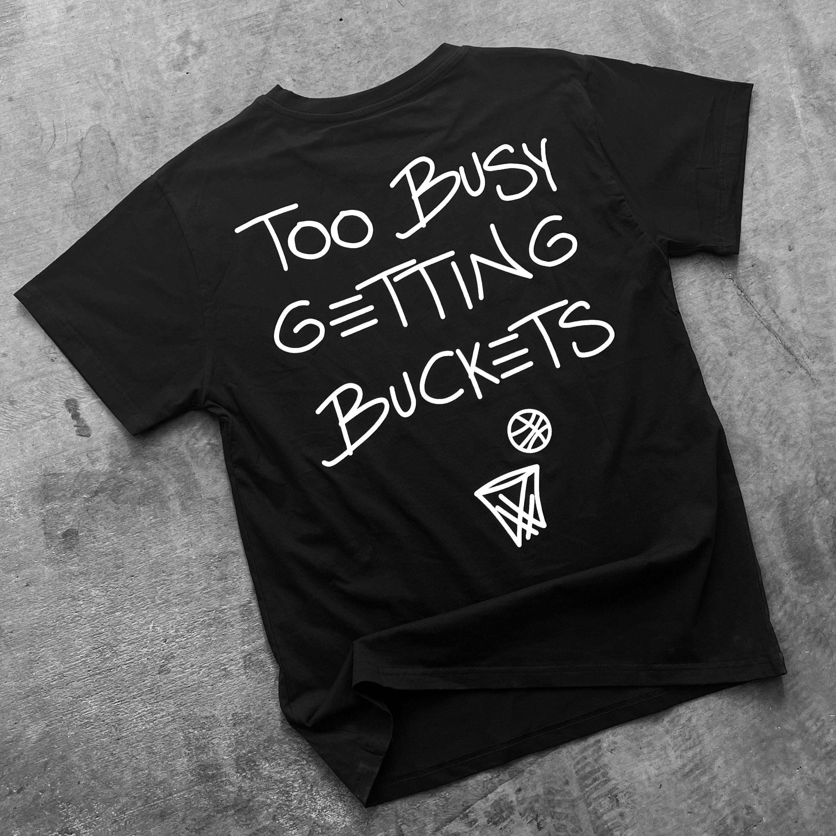 Collection of Black t-shirt with 'Too Busy Getting Buckets' text and basketball graphics on a gray surface in a gallery layout