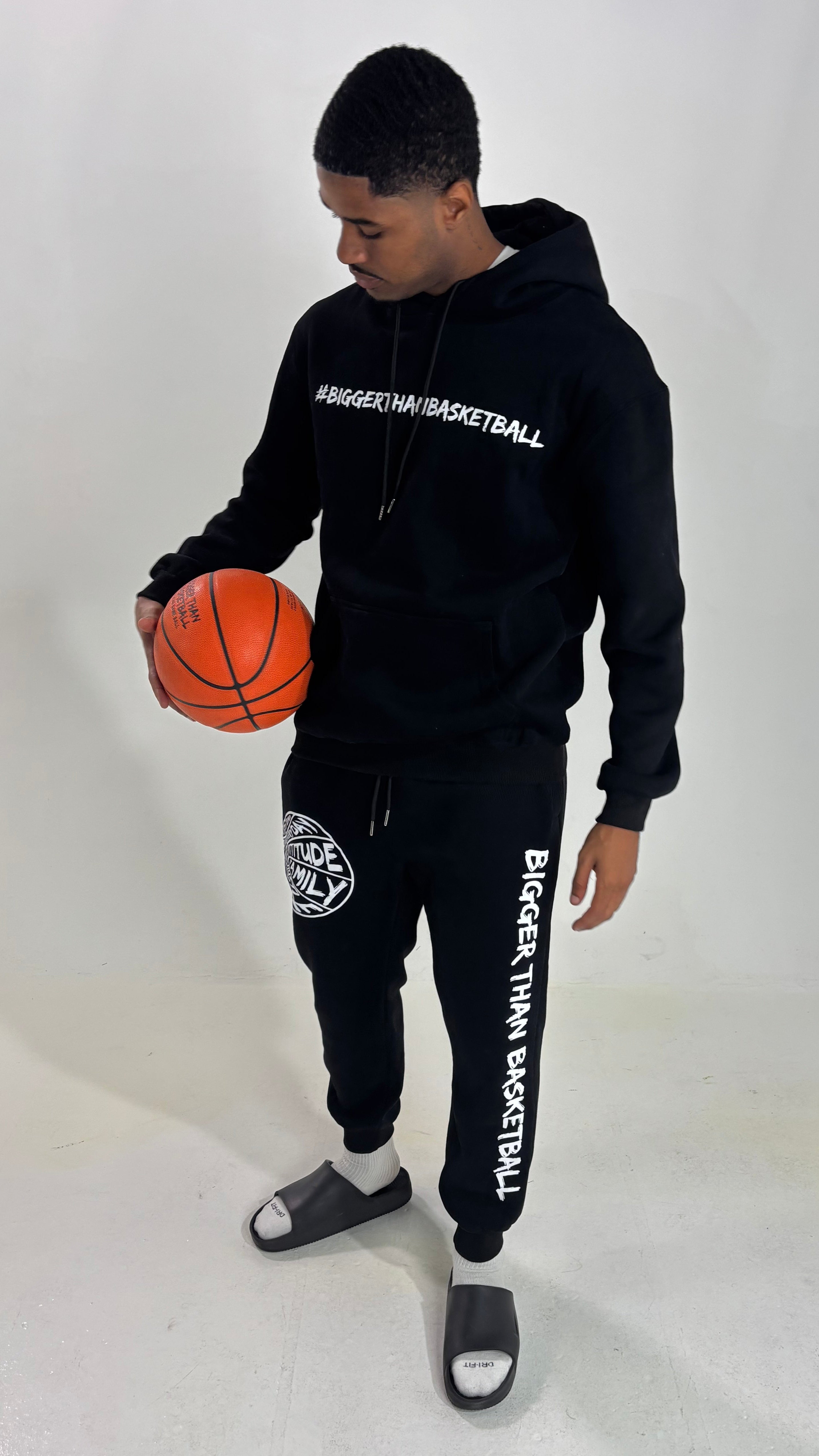 Collection of Person wearing a black 'Bigger Than Basketball' hoodie and pants holding an orange basketball on a white background in a gallery layout