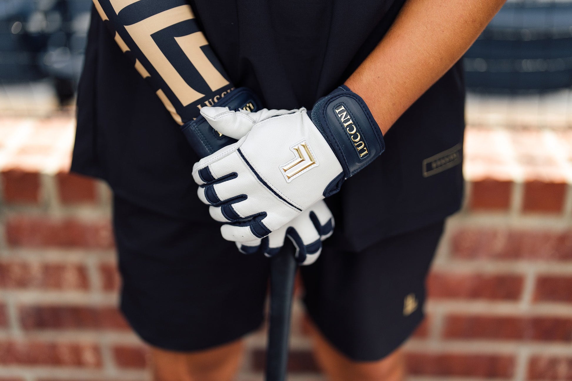 Person wearing a navy and white batting gloves holding a bat with a blurred brick wall background