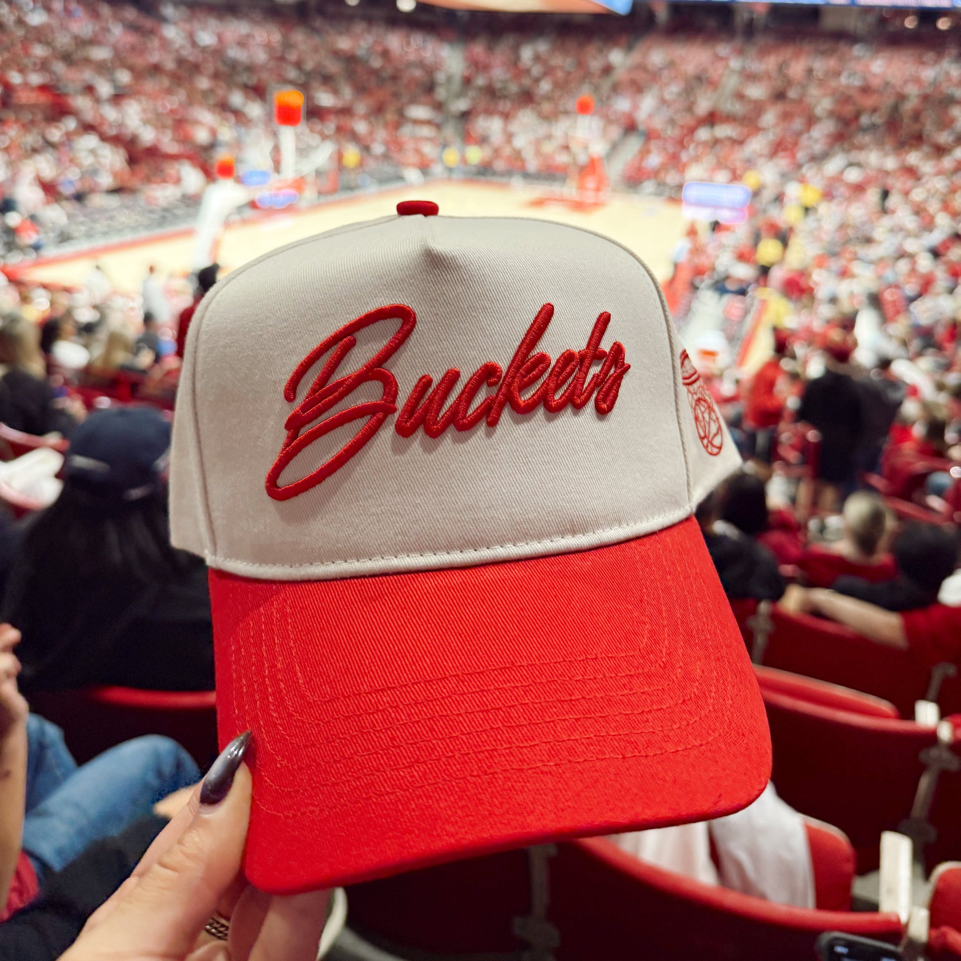 Red and beige cap with 'Buckets' text held up in front of a sports arena crowd.