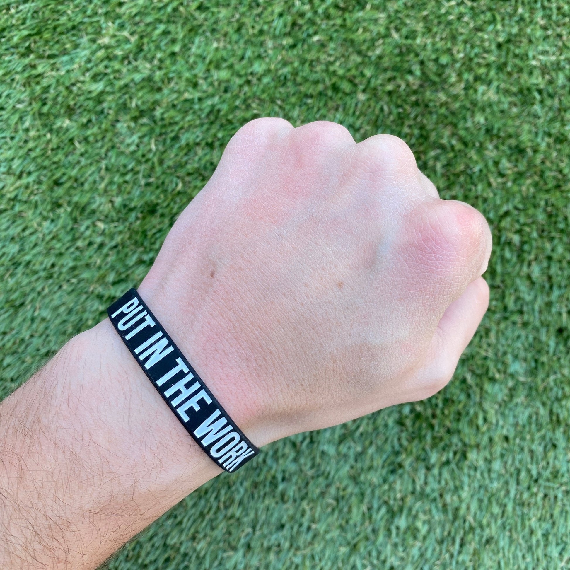 Hand wearing a black wristband with 'Put In The Work' text on a grass background