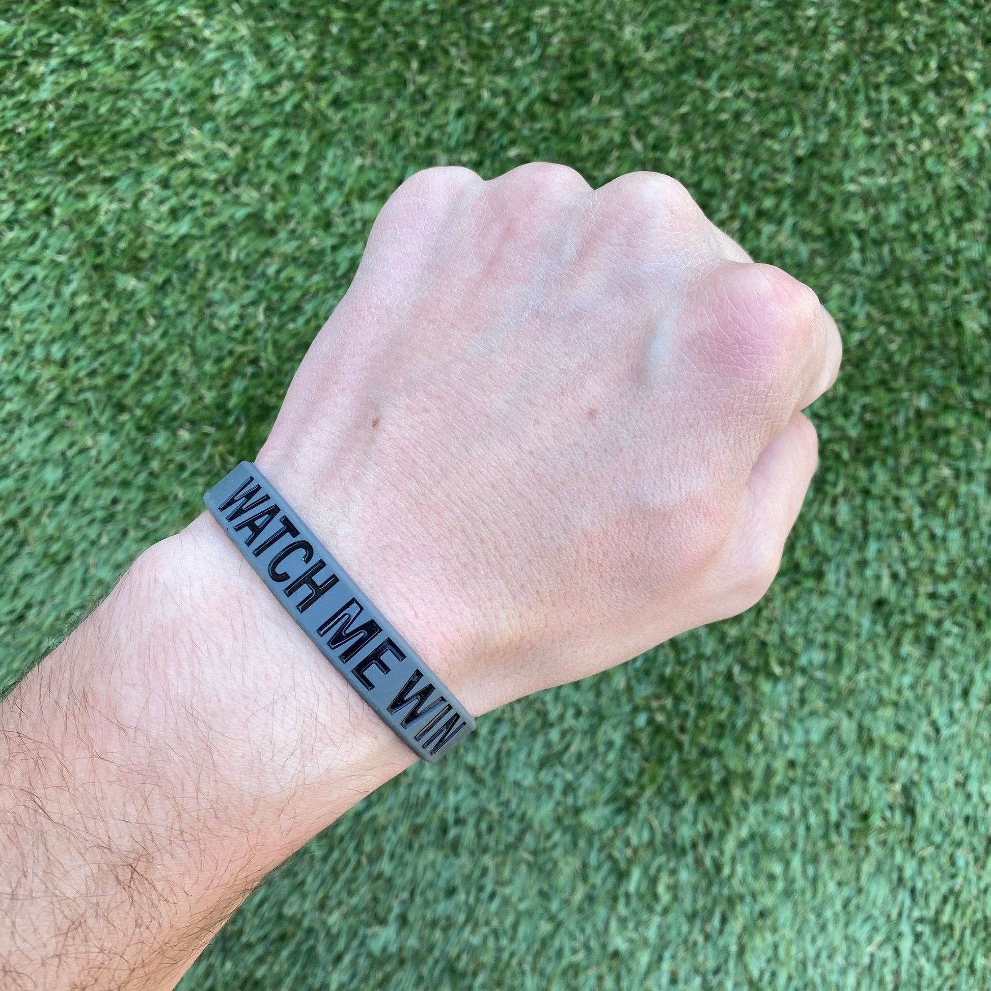 Hand wearing a black wristband with 'WATCH ME WIN' text on a grass background